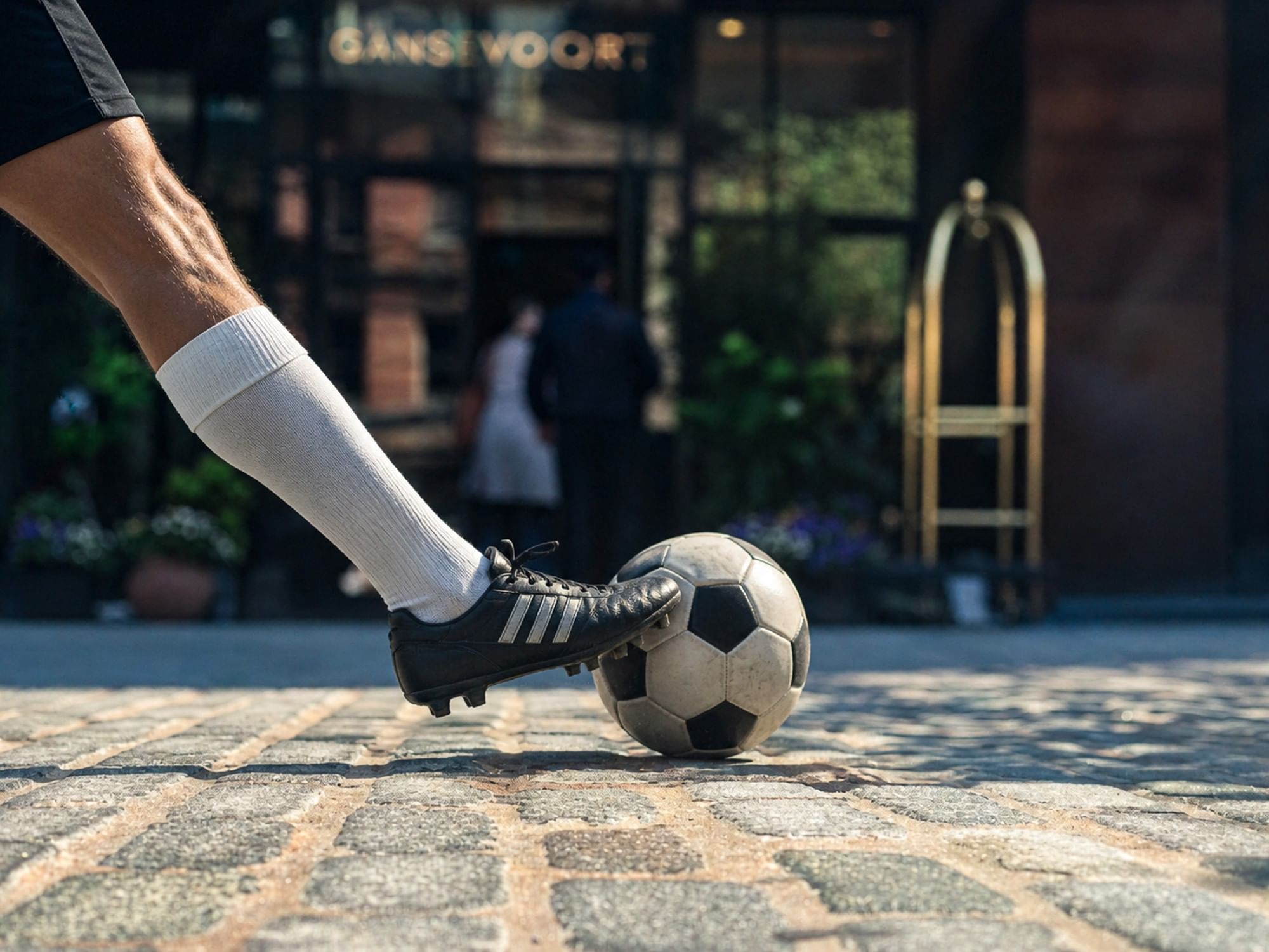 Foot kicking a soccer ball on cobblestones with blurred hotel entrance in the background.