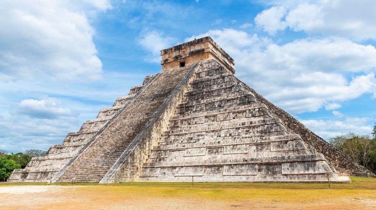Stone pyramid in a grassy field by green trees under a bright cloudy sky at Camino Real Pedregal Mexico