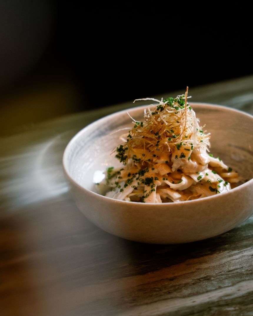 A bowl of creamy pasta topped with golden crispy onions set on a wooden table at The Kitchens