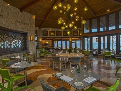 Comedor de De Cortez Grill House con sillas verdes, mesas redondas junto a los ventanales con vista al mar en Hacienda Del Mar Los Cabos