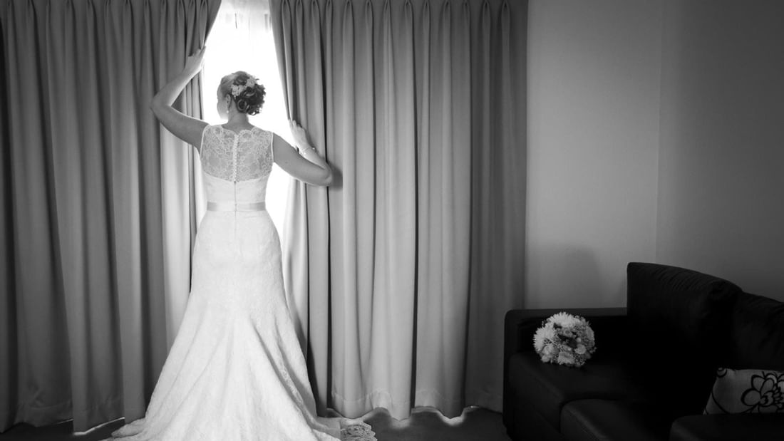 A bride in a wedding gown opens curtains, a bouquet rests on a sofa nearby in a room at Mercure Hotel Townsville