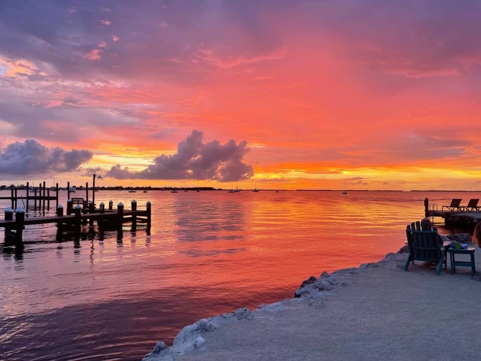 Sunset over water with a dock, chairs, and vibrant sky reflecting on the calm water surface.