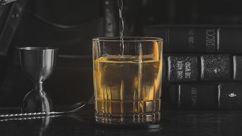 Golden whiskey with ice on a dark surface beside silver bar tools and classic books at Paramount Hotel Dubai