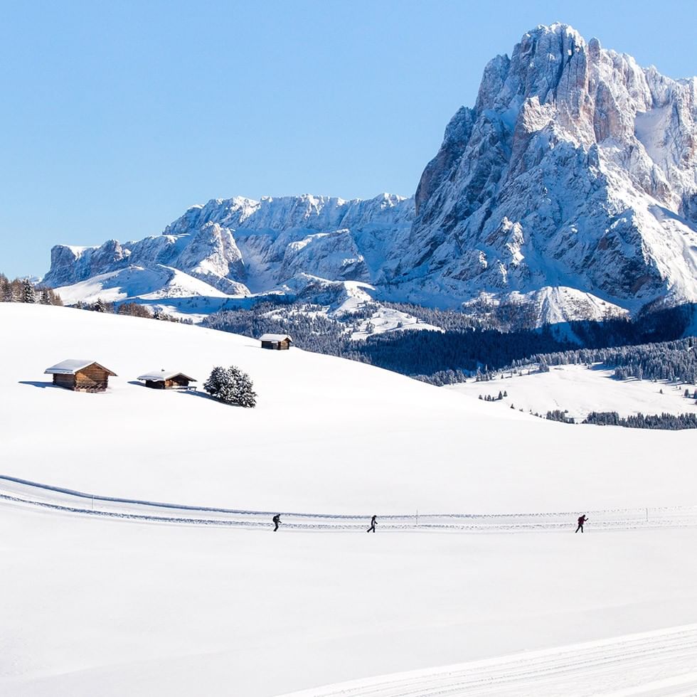 Tre persone camminano su una pista da sci innevata con montagne innevate sullo sfondo.