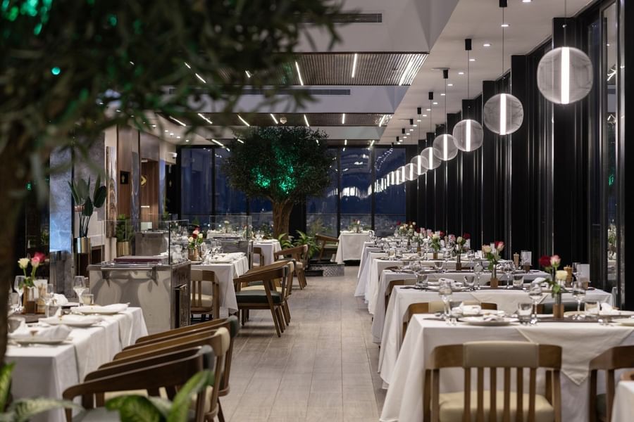 Rows of tables with white cloths by wooden chairs under globe lights in Karkan Restaurant at Warwick Al Taif Hotel 