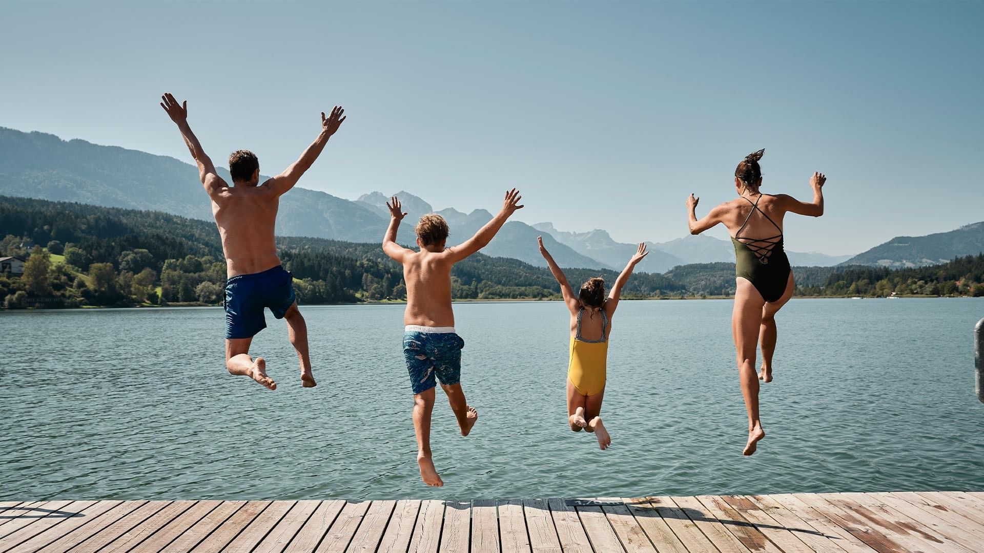 Four people jump off a wooden dock into the water with mountains in the background at Falkensteiner Resort Lake Garda