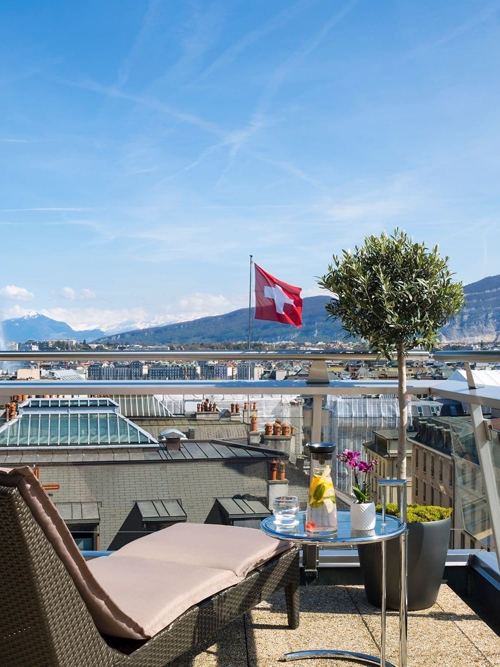 Woven lounge chair by a glass table on a rooftop terrace under a blue sky at Warwick Geneva