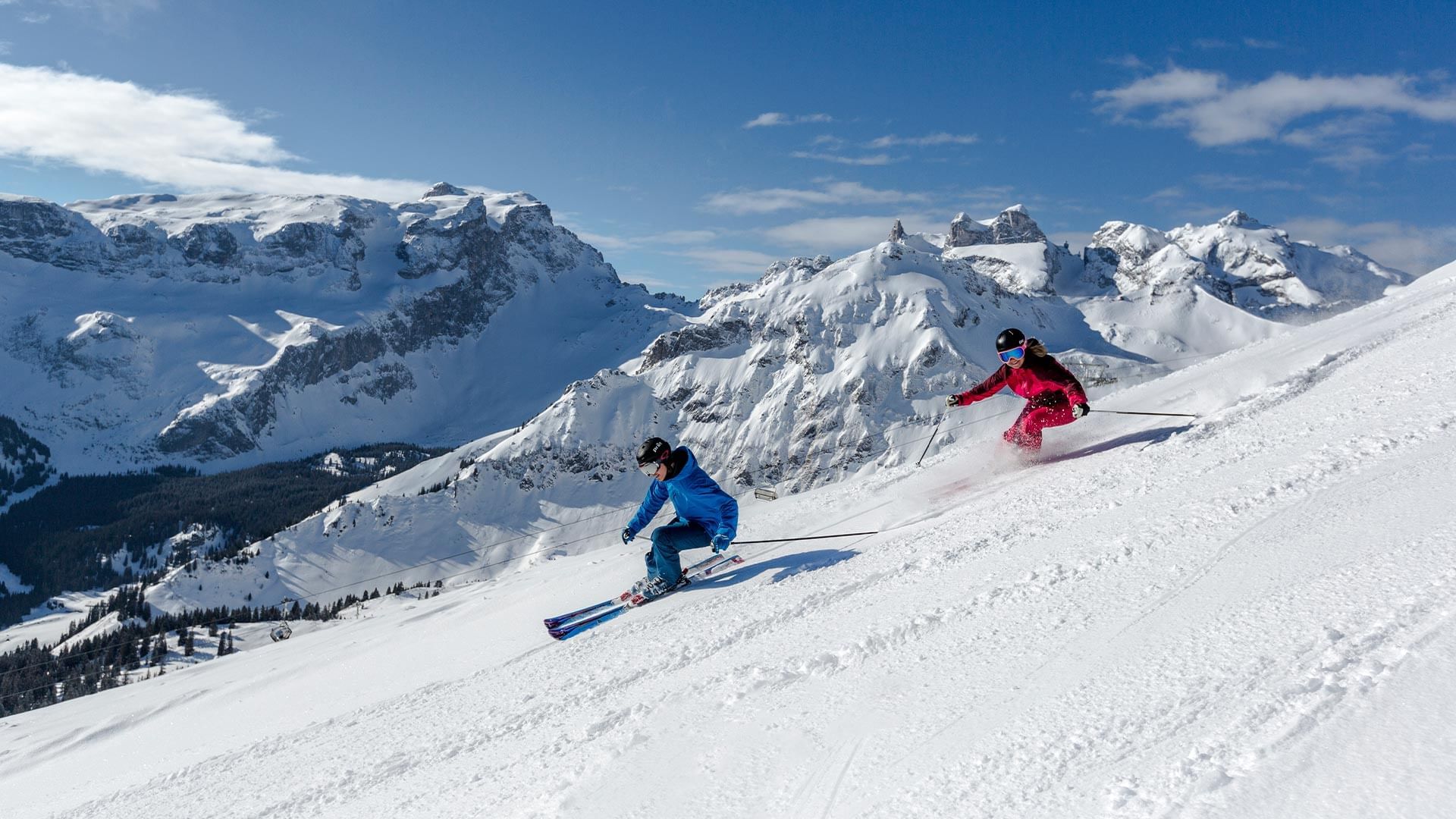 Due sciatori, uno in blu e uno in rosso, disceso su una pista da sci innevata con montagne innevate sullo sfondo.
