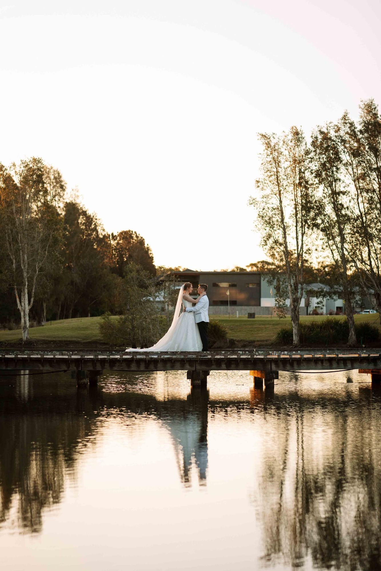 Bride and groom embrace on a bridge over a lake at sunset, with tall trees in background.