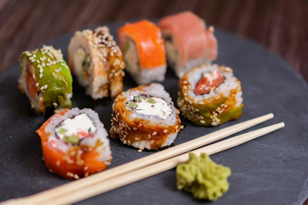 A close-up of a flat stone plate with chopsticks, wasabi, and seven unique sushi pieces featuring different fillings and sauces. 