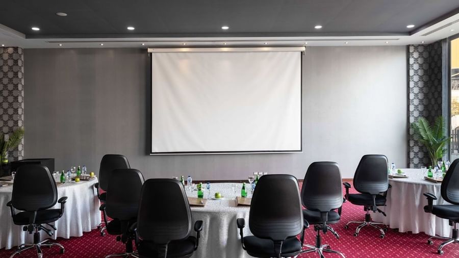 Round tables with white linens & black chairs by a projector screen on a red carpet in Dana Al Khaleej at Warwick Al Jubail