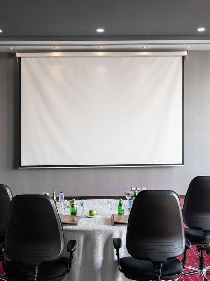 Round tables with white linens & black chairs by a projector screen on a red carpet in Dana Al Khaleej at Warwick Al Jubail
