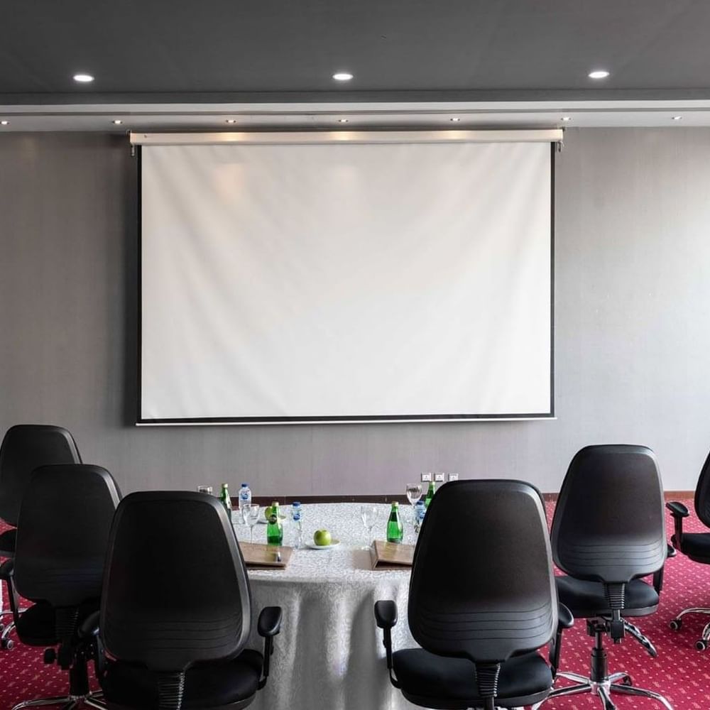 Round tables with white linens & black chairs by a projector screen on a red carpet in Dana Al Khaleej at Warwick Al Jubail