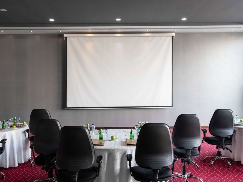 Round tables with white linens & black chairs by a projector screen on a red carpet in Dana Al Khaleej at Warwick Al Jubail