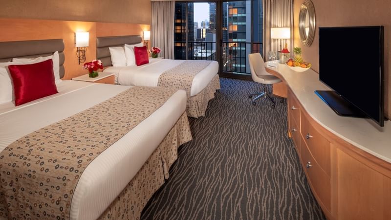 Two beds, a desk, a chair, a balcony, and a TV are in a hotel room.