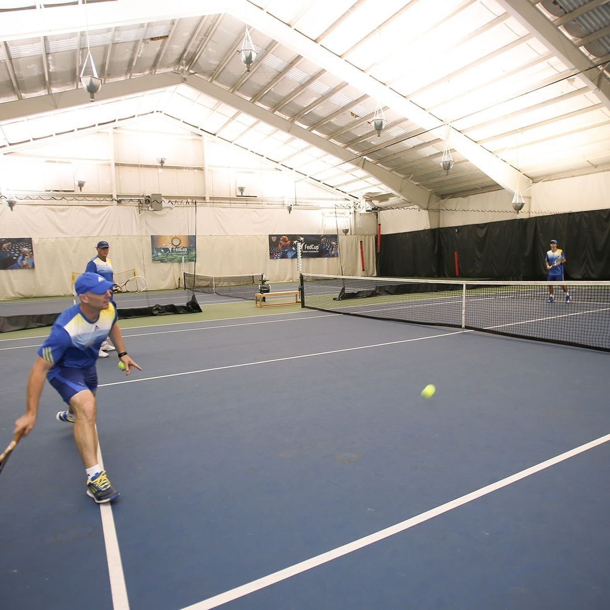 Players in blue uniforms engaged in a cardio tennis and point play session on an indoor court with white ceiling.