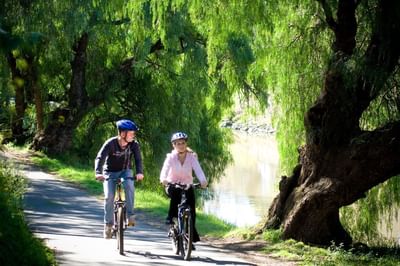 Two people riding bikes on a sunny, tree-lined path beside the Yarra River near Amora Herencia Riverwalk Melbourne