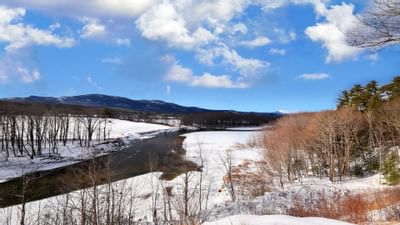 A river flows through a snowy landscape near the Fox Ridge Resort with bare trees and distant mountains under a cloudy sky.