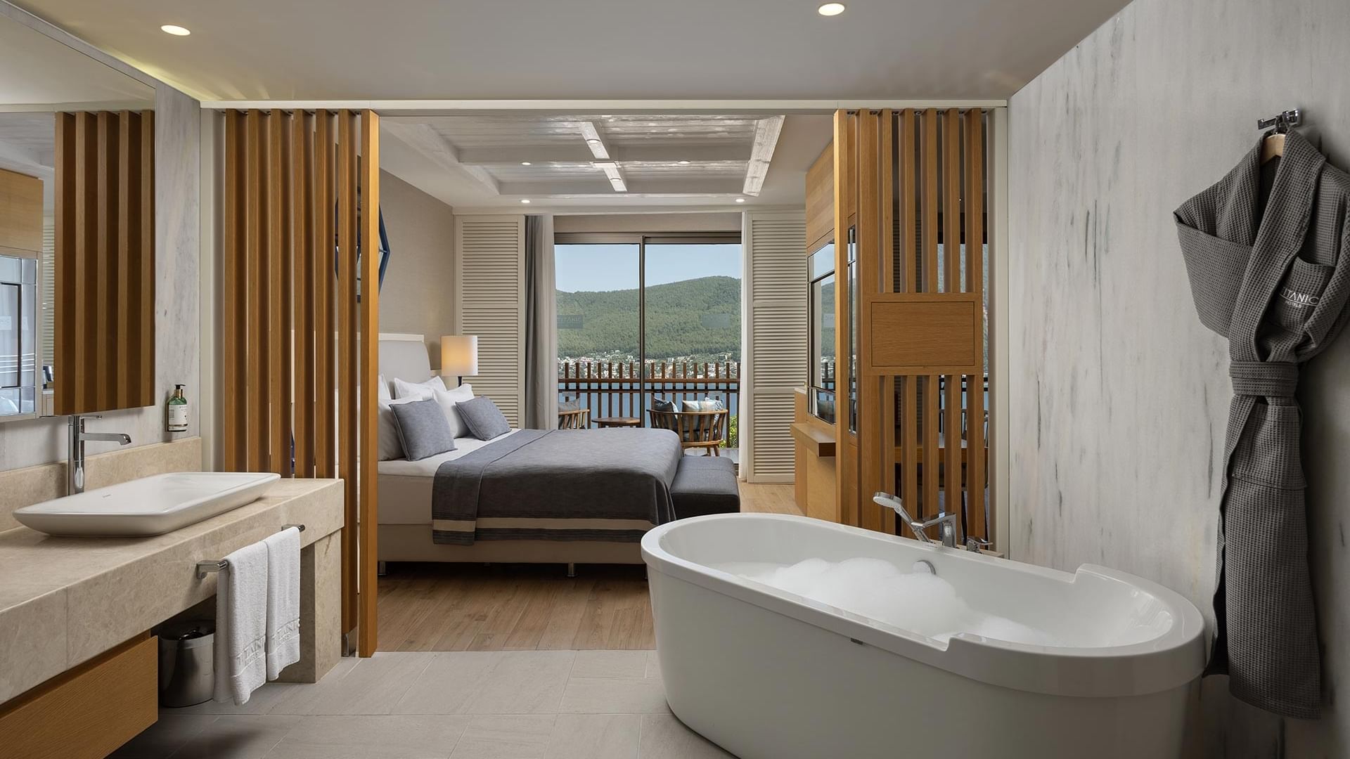 Modern bathroom with large bathtub, sink, and open bedroom with balcony and mountain view at Titanic Luxury Collection Bodrum..