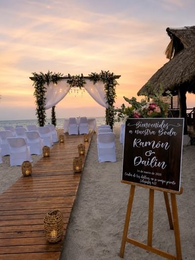 Beachfront wedding ceremony setup with floral arch, white chairs, and sunset over ocean at Playa Blanca Beach Resort