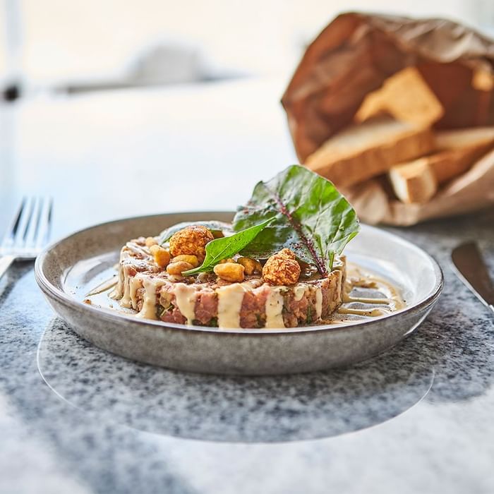 Piatto di tartare di carne con verdure e salsa, accanto a una borsa di pane su una superficie in pietra.