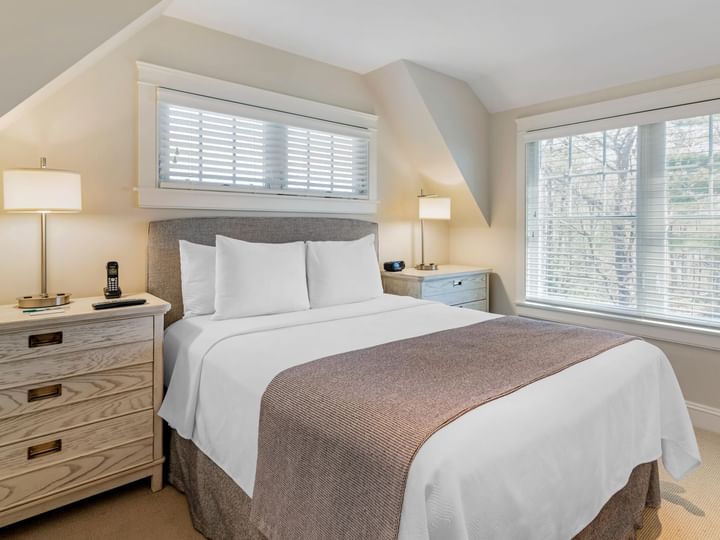 A neatly made bed with white linens and a brown throw blanket in a room with beige walls, two windows, and a wooden dresser.