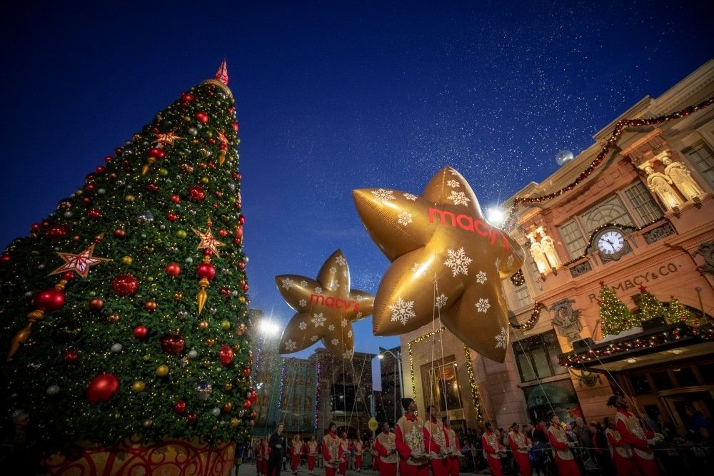 At twilight, a large Christmas tree sparkles next to floating gold star balloons carried by a team of balloon wranglers at Universal Studios Florida.