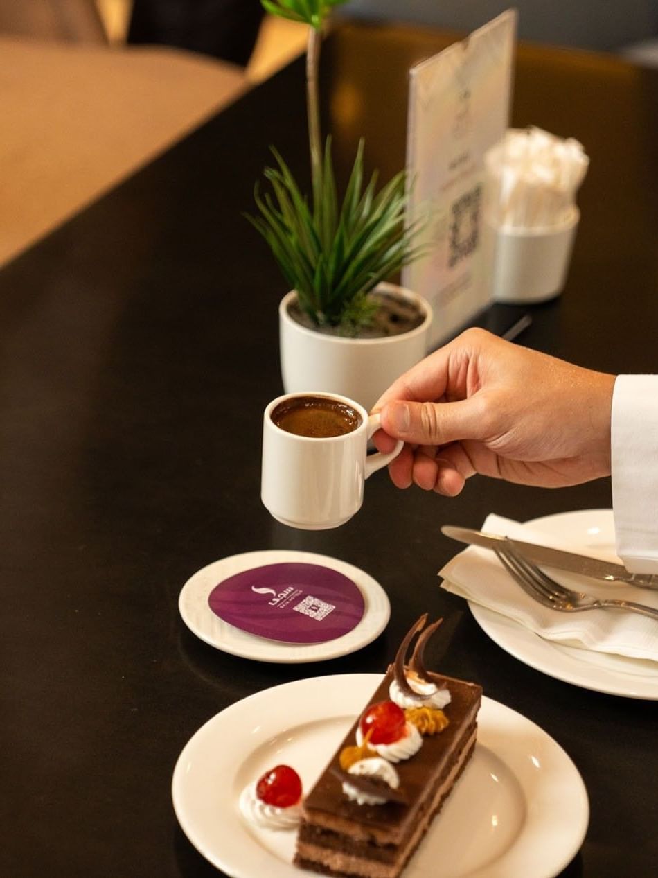 Close-up of a hand holding a cup of Arabic coffee next to a slice of chocolate cake at Saja Warwick Madinah