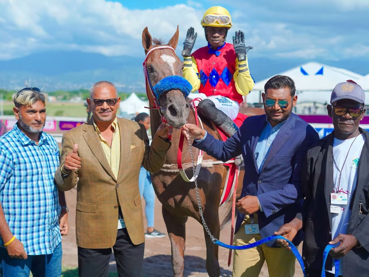 Jockey holding hands up beside owners posing with a winning brown racehorse near Jamaica Pegasus Hotel