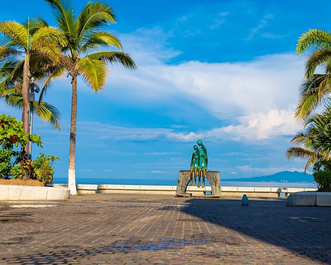 La escultura 'La Soledad' en el malecón de Puerto Vallarta, que muestra dos figuras de bronce estilizadas en un banco de piedra frente al mar y la Bahía de Banderas, con la zona de playa de Los Muertos al fondo bajo un cielo azul.