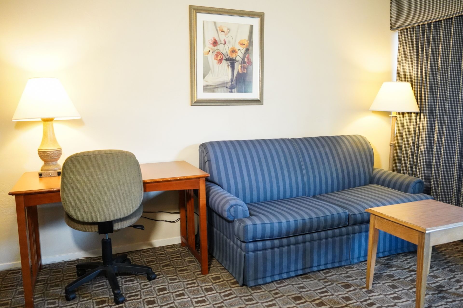 A hotel room living area with a blue striped sofa, wooden desk, office chair, coffee table, and two lamps.