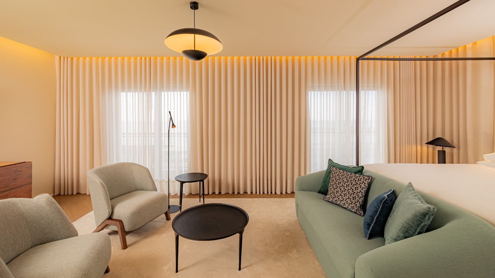Quarto de hotel com cama, sofá, mesa, cadeiras, cortinas e luminárias.