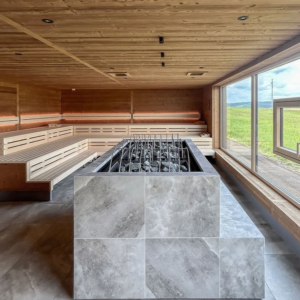 Wooden sauna with large windows, wooden benches, and a central stone stove with rocks.