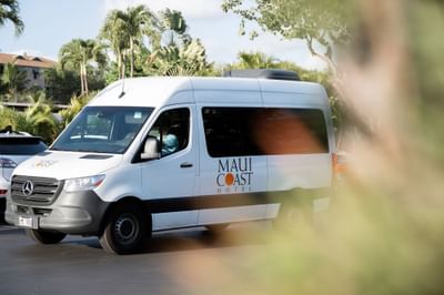 White Mercedes-Benz shuttle van with the Maui Coast Hotel logo, parked among palm trees at the hotel's entrance