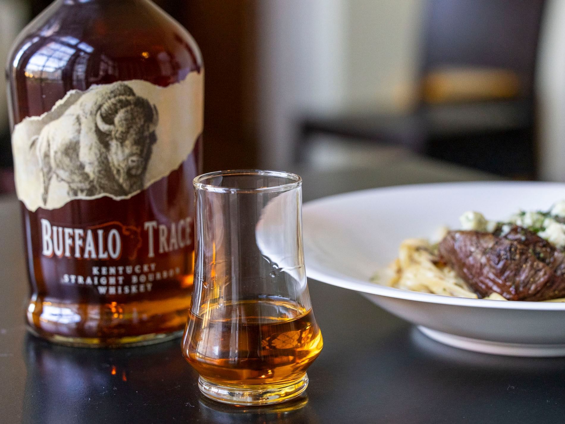 Whiskey glass in the foreground with a bottle of Buffalo Trace and steak pasta in the restaurant at The Ashbrook Hotel