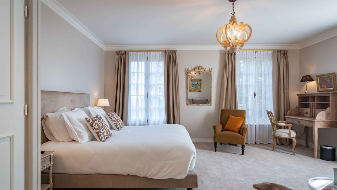 Chambre luxueuse avec lit, chaises, bureau, miroir, lustre et deux fenêtres avec rideaux.