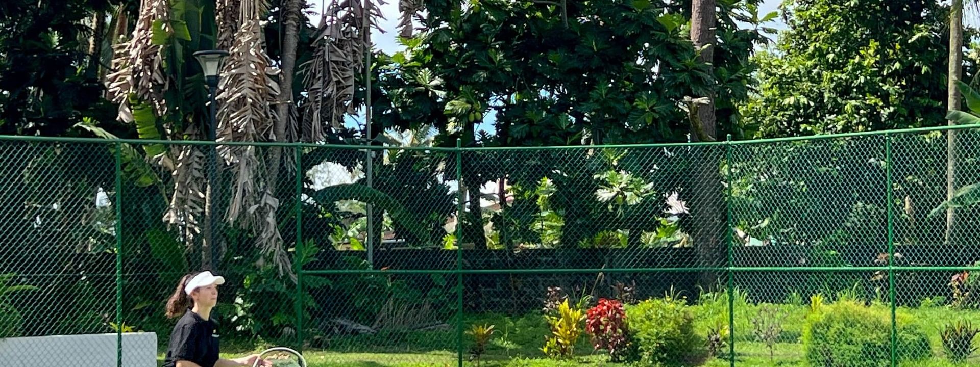 Young woman playing tennis by a chain link fence under a sunny sky at The Naviti Resort in Korolevu
