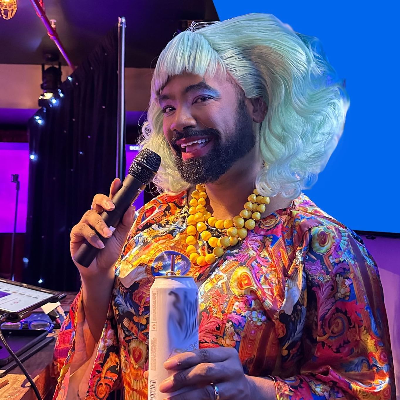 A performer in a colorful, patterned garment holds a microphone, with a large necklace in a Karaoke Night at The Gaythering