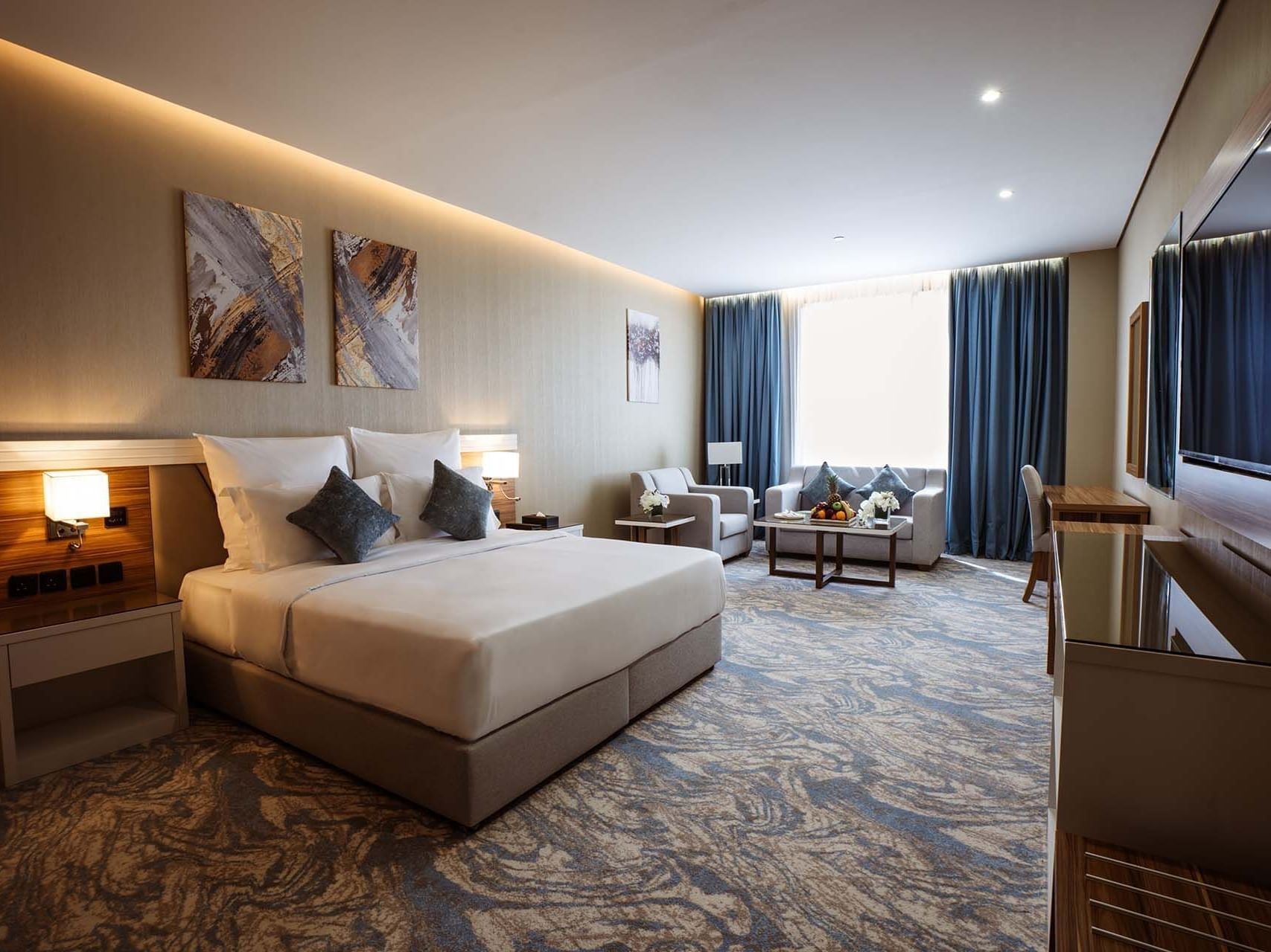 Large bed in Deluxe Room with paintings and a sofa under blue curtains by a window at Cantonal Hotel by Warwick Riyadh