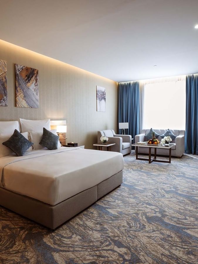 Large bed in Deluxe Room with paintings and a sofa under blue curtains by a window at Cantonal Hotel by Warwick Riyadh