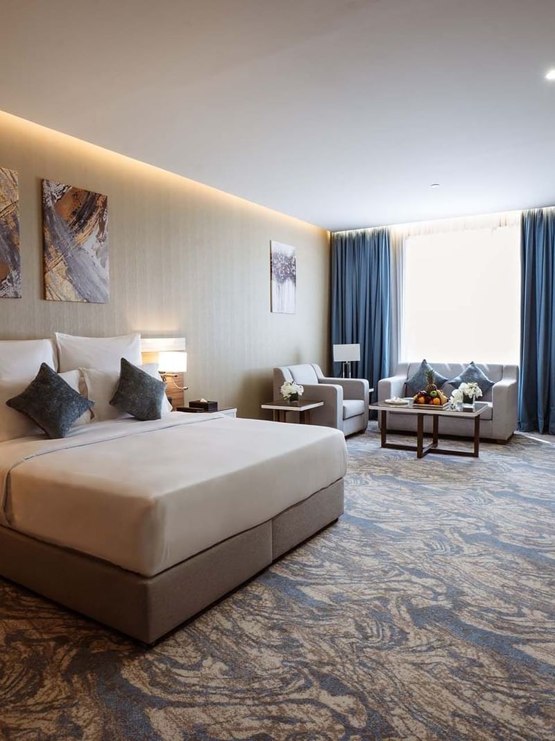 Large bed in Deluxe Room with paintings and a sofa under blue curtains by a window at Cantonal Hotel by Warwick Riyadh