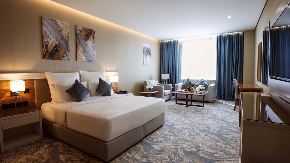 Large bed in Deluxe Room with paintings and a sofa under blue curtains by a window at Cantonal Hotel by Warwick Riyadh
