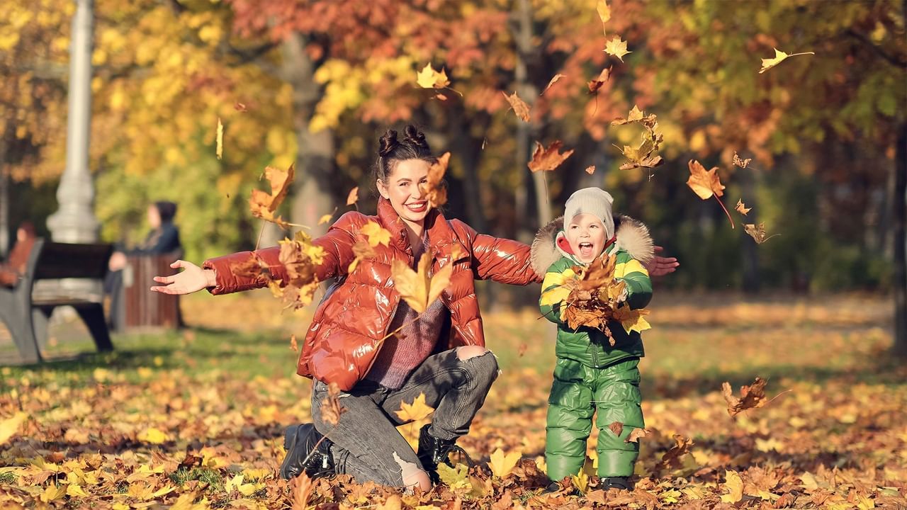 Mother and child joyfully tossing colorful autumn leaves in a park with vibrant fall foliage in the background