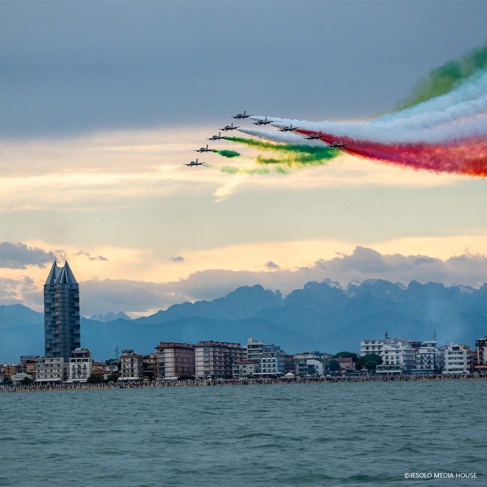 Aerei militari in formazione sopra una spiaggia affollata e una città costiera all'alba.