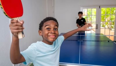 Smiling young boy holding a red paddle and white ball by a blue ping pong table indoors at Worthing Court Apartment