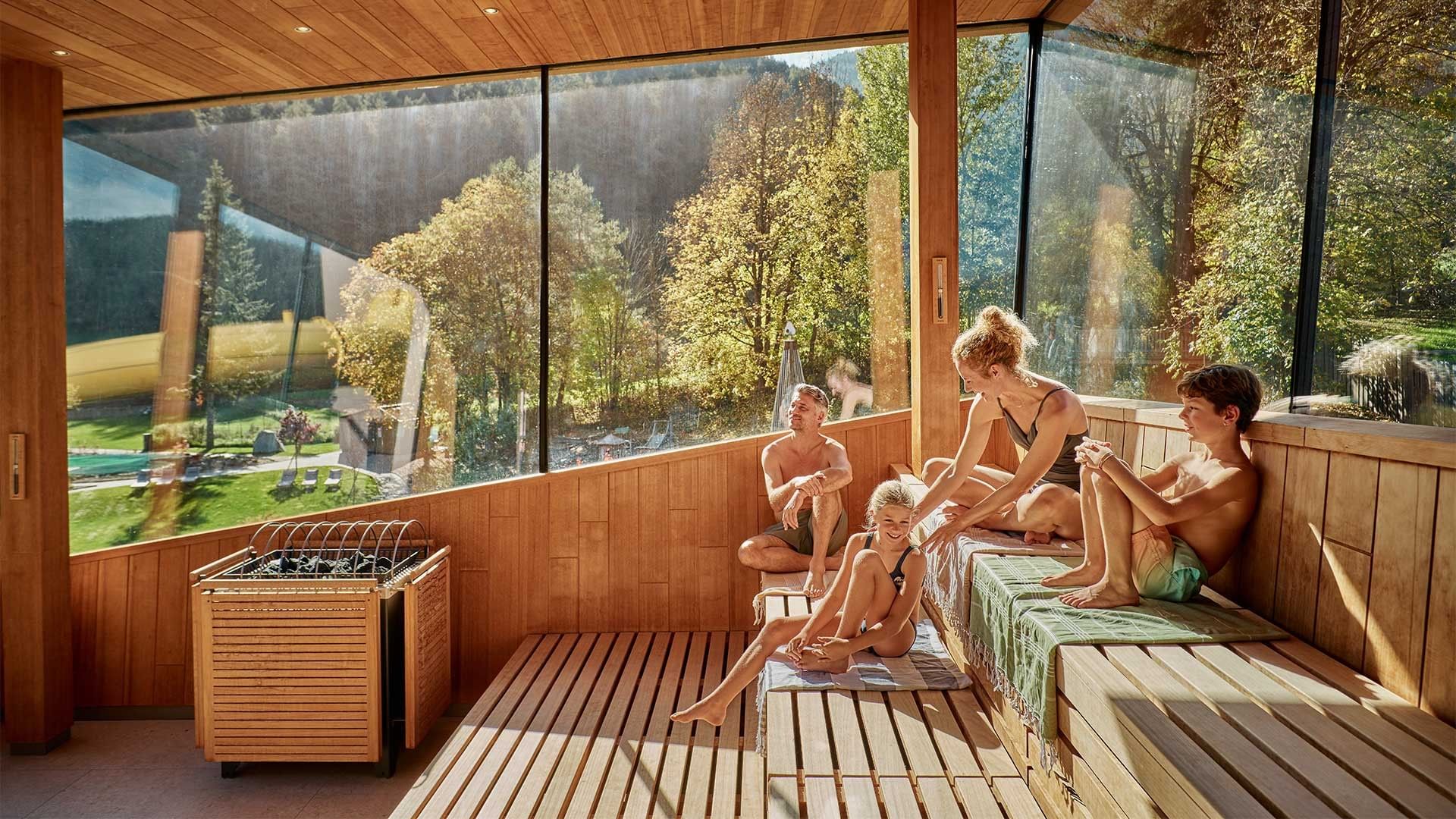 Famiglia rilassata in una sauna con grandi finestre che si affacciano su un paesaggio verde.