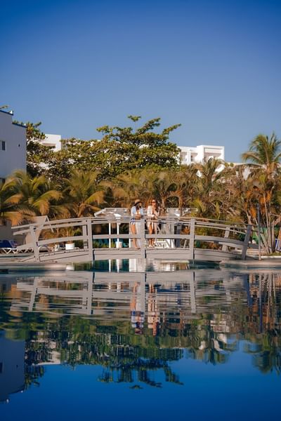 Two guests on a white bridge over a pool surrounded by palm trees at Playa Blanca Beach Resort