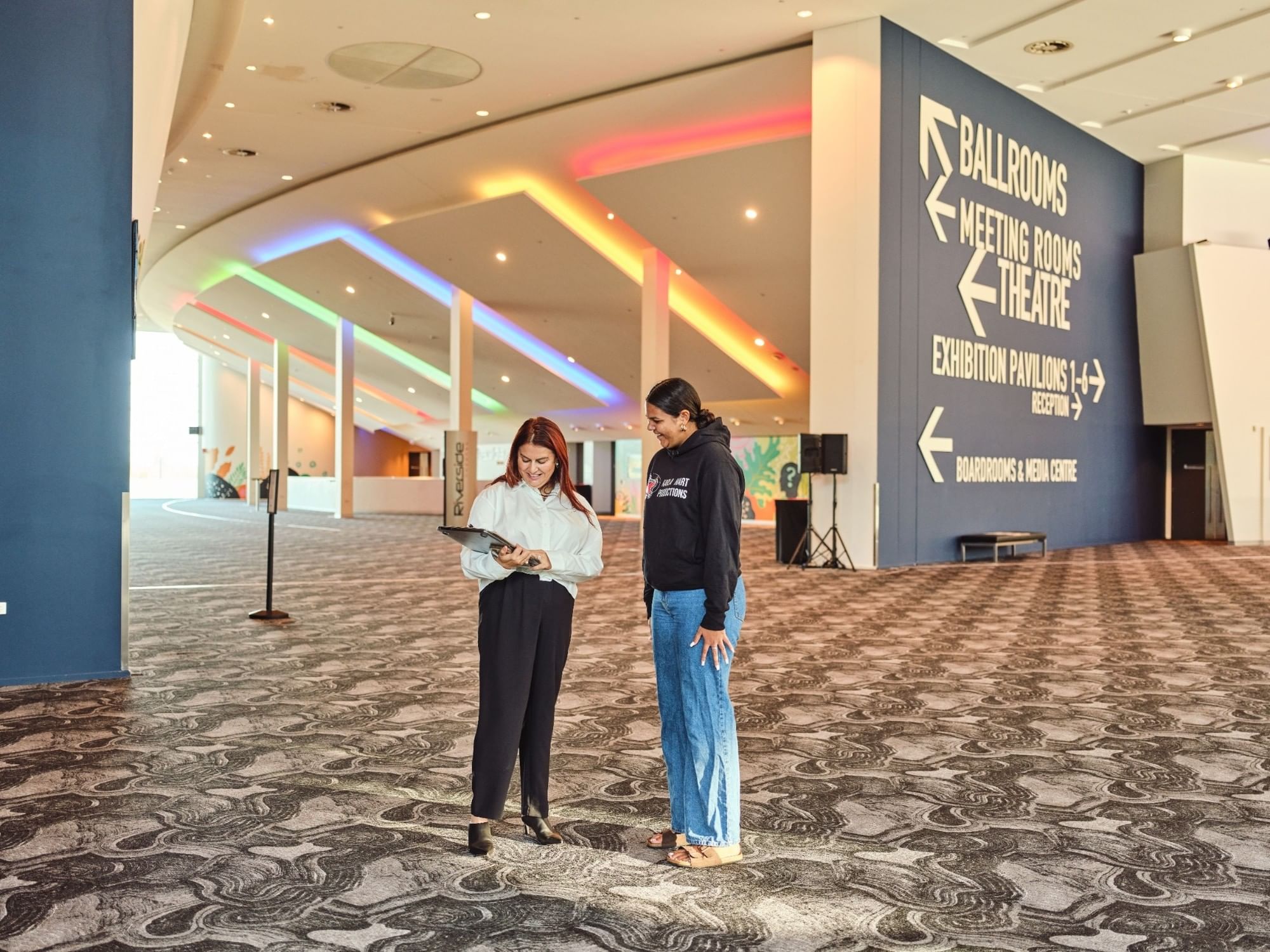 Two people reviewing materials in Perth Convention and Exhibition Centre foyer with colorful ceiling lights and directional signage.