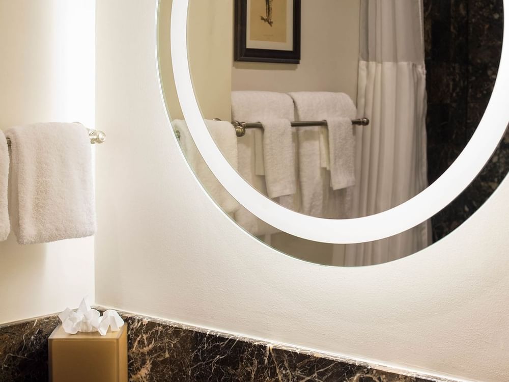 Round glowing mirror by white towels on a rack surrounding a marble vanity in Twin Room at Warwick San Francisco