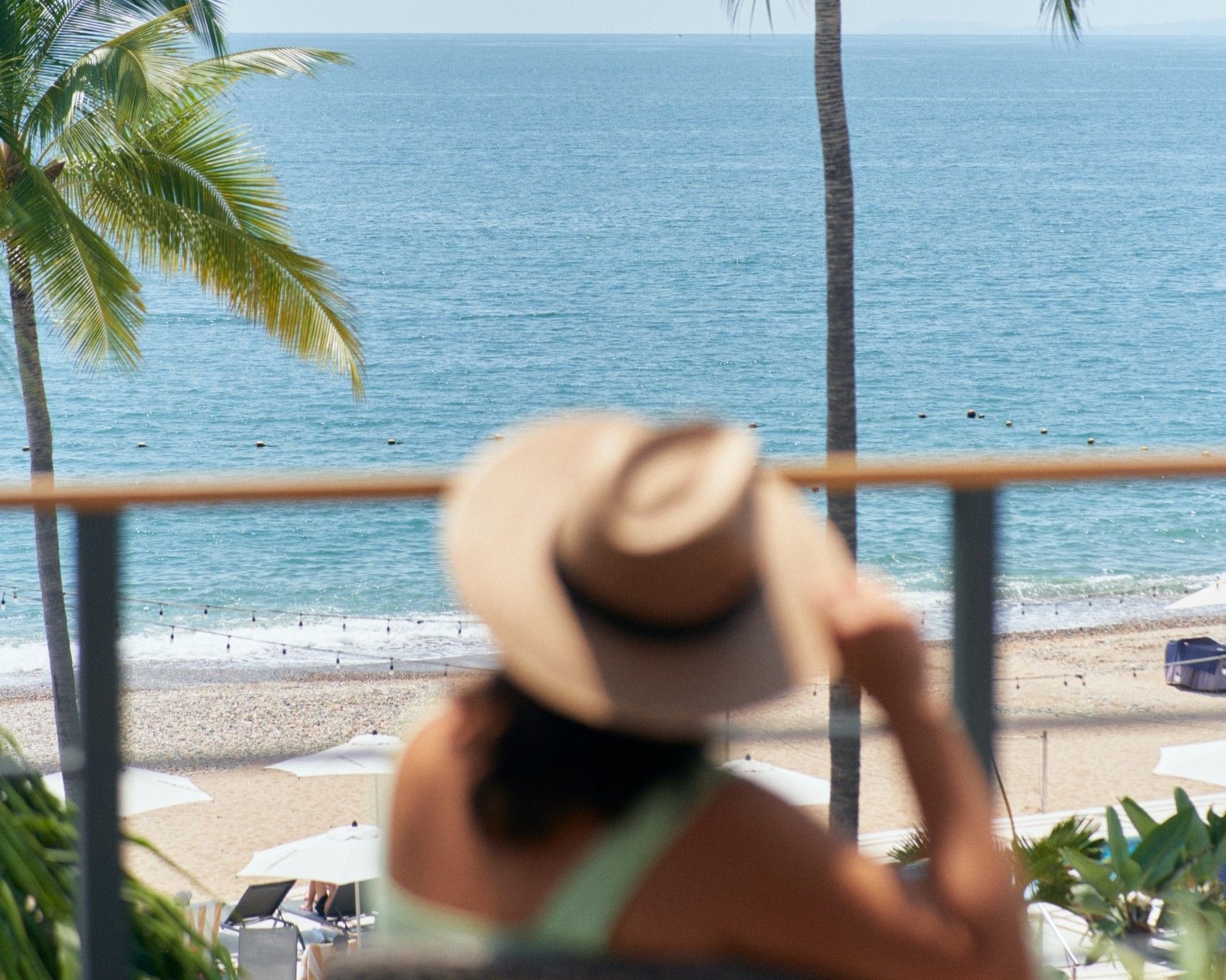 A woman wearing a sun hat looks out from a balcony at the Buenaventura Grand Hotel, overlooking a sunny beach with white umbrellas and the blue ocean in Puerto Vallarta.